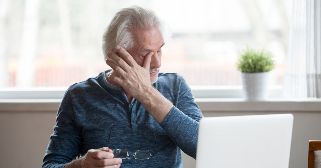 A person wearing a long-sleeved blue shirt is sitting in front of a computer and rubbing their eyes.