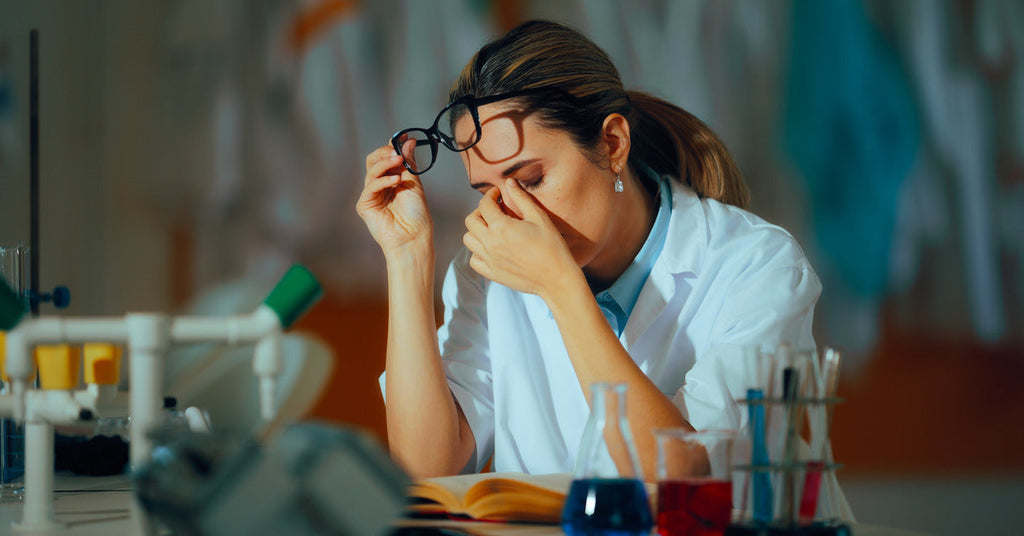 A professional in a lab coat is removing their glasses and rubbing their eyes while leaning over a book on a table.