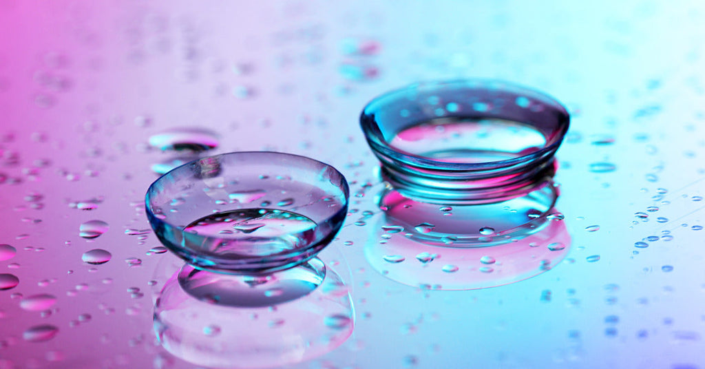 Two contact lenses are sitting on a shiny pink and blue surface. The lenses are surrounded by water droplets.