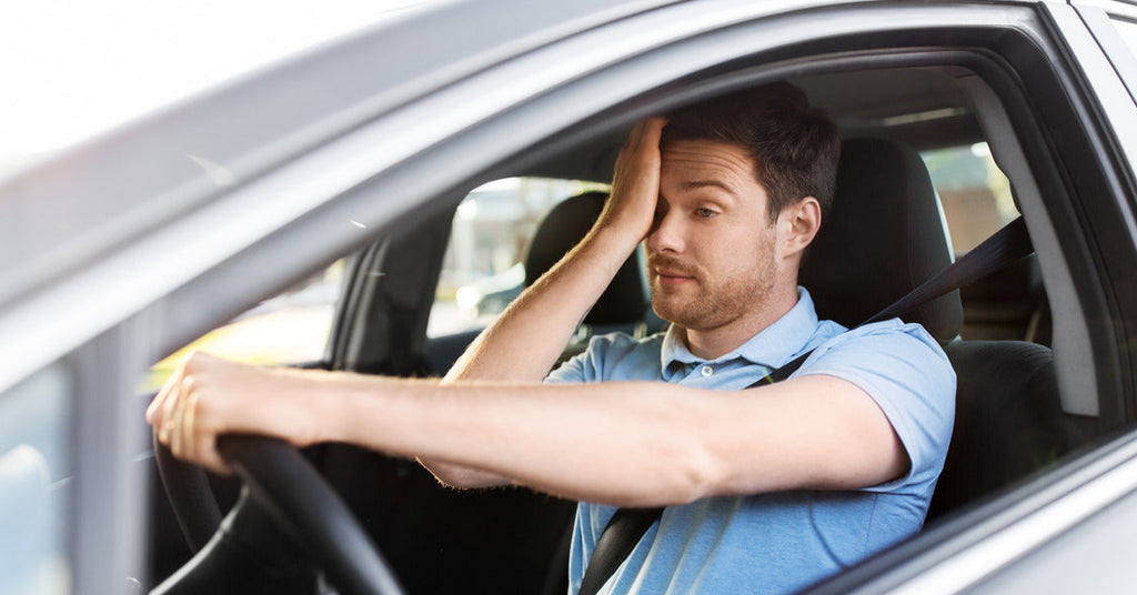 A person wearing a blue T-shirt is sitting behind the steering wheel of their car while rubbing their right eye and looking tired.