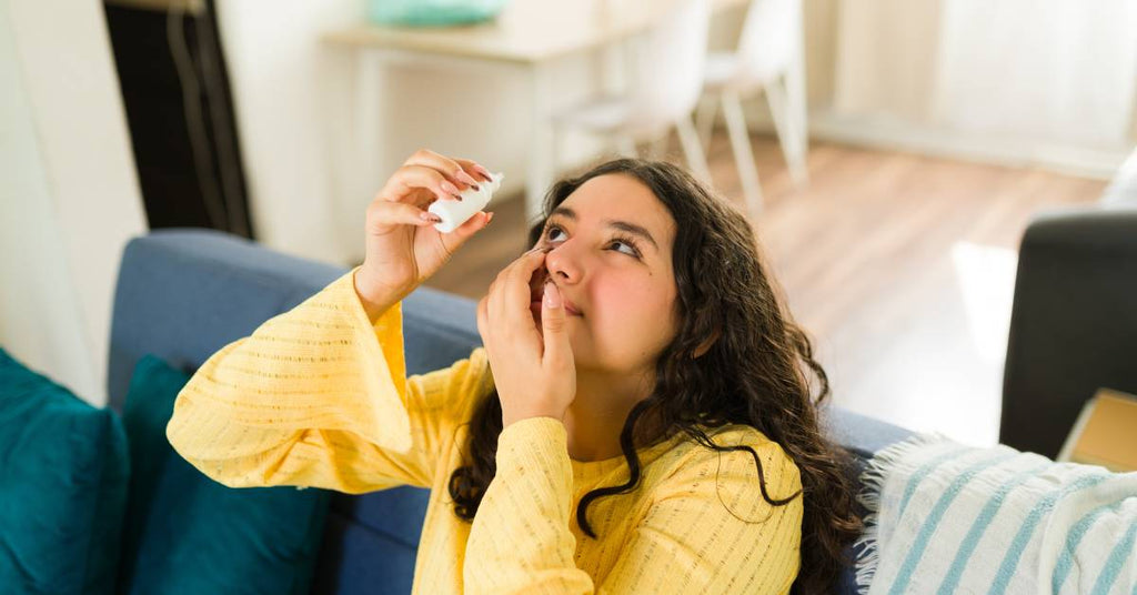 A teenage girl in a yellow sweater sits on a couch, leaning her head back to use eye drops.